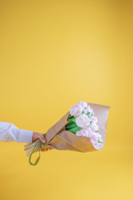 A hand extending from the left, clad in a white shirt, holding a bouquet of fresh, pastel pink tulips wrapped in brown craft paper, tied with a green ribbon. The tulips are in full bloom with soft, de