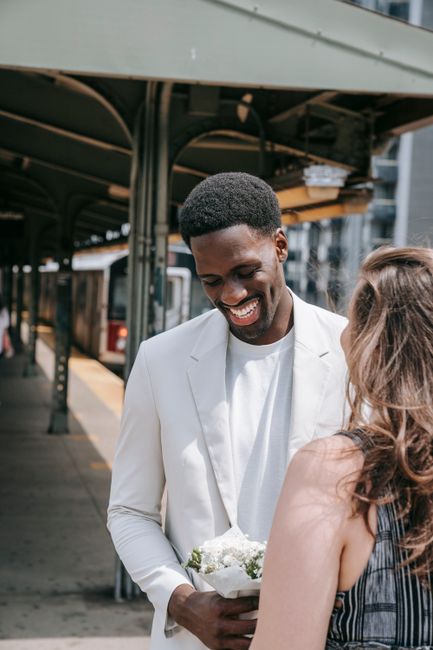A smiling man with dark skin, wearing a white blazer over a white shirt, holds a bouquet of white flowers wrapped in light paper, standing at a train station platform. The woman in front of him has li