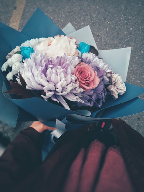 A hand holding a bouquet of fresh flowers wrapped in blue and white paper, featuring a large white chrysanthemum, a pale pink rose, light purple lisianthus, and smaller white blossoms. The bouquet is 